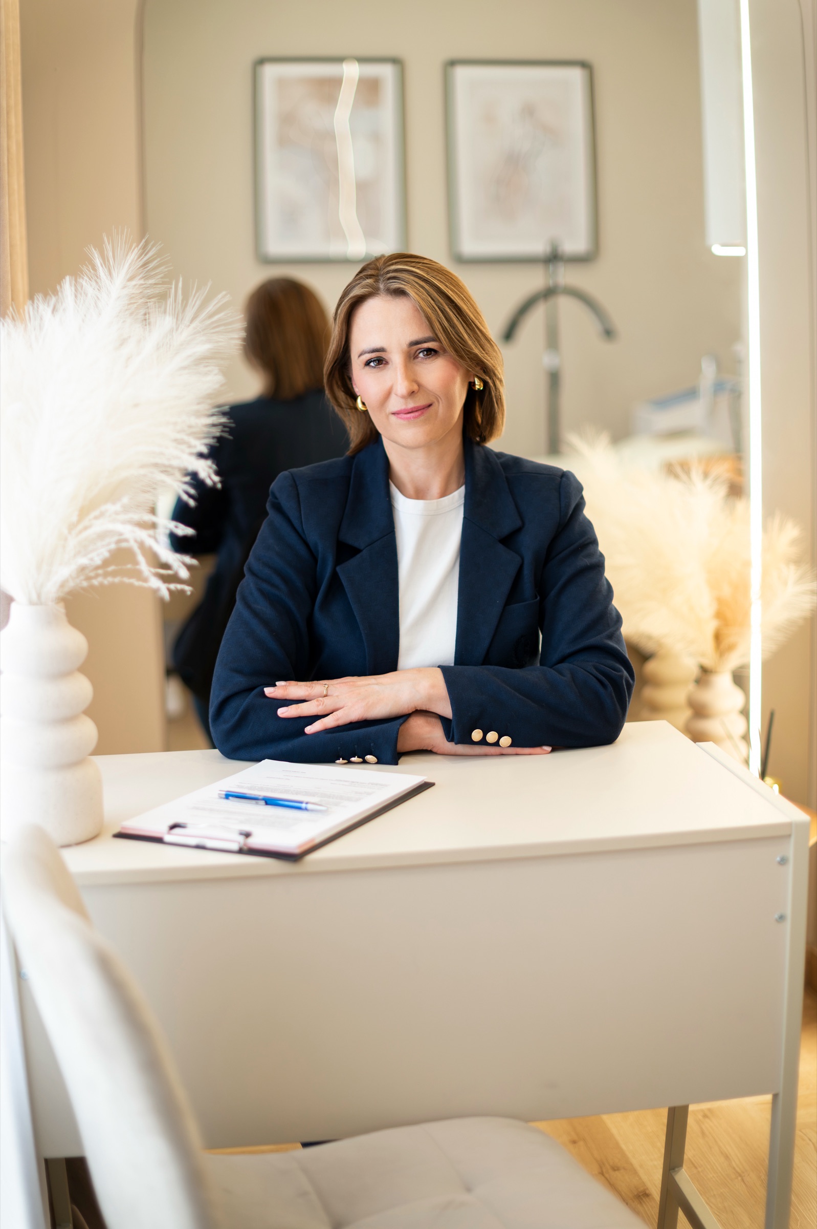 Dorota Bazan przy białym biurku konsultacyjnym z kartą pacjenta, w tle wazony z pampasami